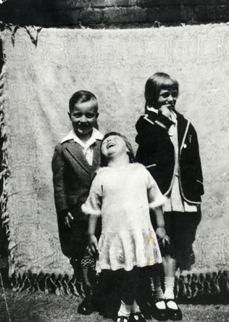 Photograph showing three children standing in front of a blanket suspended behind them; on the left is a boy, aged approximately six years, wearing an open-necked shirt, a jacket, long socks and dark shoes; on the right is a girl, aged approximately nine years, wearing a dark blazer with a light border, a pleated skirt, socks and button shoes; in front of the other two is a child, aged approximately four years, wearing a light-coloured dress, socks and sandals; they have been identified as being in Blackhall
