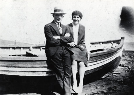 Photograph of Jenny Petty and Friend, showing a man, dressed in suit and wearing a Trilby hat, and a woman, wearing a suit and a beret, sitting on the edge of a rowing boat, possibly on Blackhall beach,1930