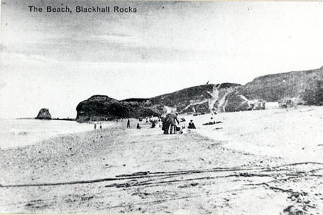 Postcard photograph entitled The Beach, Blackhall Rocks, showing the beach looking south with the rocks in the distance, and a number of people on the beach in the middle and far distance; in the near distance a number of chains or ropes are lying across the beach