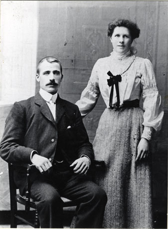 Photograph, taken in a photographer's studio, showing a man dressed in a suit with stiff collar and watch chain, sitting in a chair, and a woman, dressed in a blouse with tucks and embroidery and in a long skirt, standing beside him: they are identified as Mr. P Ward,Sinker, and his wife