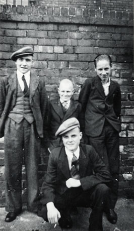 Photograph of four young men in front of a high wall, identified as Wilf Riley and friends of Blackhall; one young man is dressed in a suit, tie and cap; another in a suit and tie; a third in a suit without a tie; the fourth, crouching on the ground, is wearing a suit, tie, and cap and is smoking a cigarette