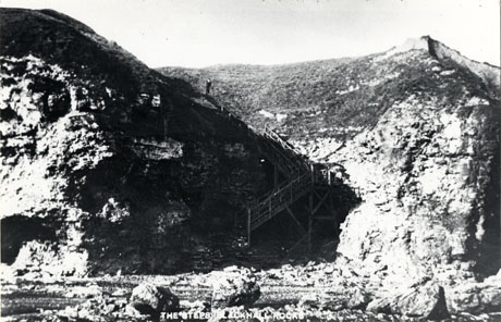 Postcard photograph, entitled The Steps, Blackhall Rocks, showing the rocks at Blackhall fronting the sea and a set of steps, possibly made of wood, coming down the face of the rocks with a small figure at the top of the steps