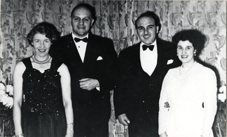 Photograph of two men and two women in evening dress, photographed in front of large and luxurious curtains, possibly in an hotel; the men are wearing dinner jackets and one woman a dark evening dress with bare arms and the other a light evening dress with long sleeves; they are described as Leo and Doris Pieroni and Mr. and Mrs. Magiore
