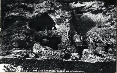 Postcard photograph, entitled The Cave Entrance Blackhall Rocks, showing the front of the rocks facing the sea and the entrances to two caves, with two indistinct figures standing at the mouth of the higher of the two caves