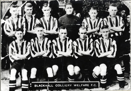 Photograph showing ten men in a strip of dark shorts and striped shirts and a man in a dark jersey; a number of indistinct faces can be seen behind the group; the footballers have been identified as members of the Blackhall Welfare Football Team