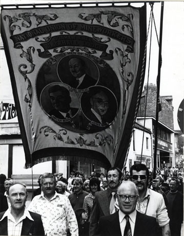 Photograph of people walking along North Road in Durham City ,towards Framwellgate Bridge, during the Miners' Gala; at the front of the photograph are a number of Blackhall Lodge Officials, with the Blackhall Lodge banner, on which there are portraits of Earl Attlee, Aneurin Bevan,and Arthur Horner