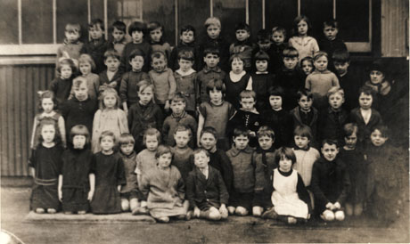 Photograph showing a group of fifty two children, aged approximately seven years, posed in front of a building of corrugated iron, with, presumably, their female teacher standing to the left of the children