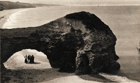 Photograph, looking north, of the outcrop of rock at Blackhall, showing a number of people standing under the rock on the beach with the coastline curving round in the distance; the people under the rock are indistinct but one appears to be wearing a long skirt