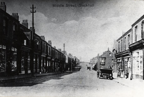 Postcard photograph entitled Middle Street, Blackhall, showing shops on either side of the road running away from the camera into the distance; the names on the shops are indistinct; a small number of indistinct figures may be seen; an open car can be seen on the right- hand side of the picture, and another vehicle in the distance