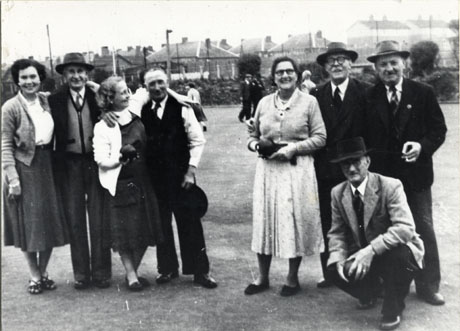 Photograph of a bowling green with, in the distance, houses, and a number of people walking on the bowling green; in the foreground, in close-up, are five men and three women; two women are holding bowling balls and one man is squatting down and holding a bowling ball; all the figures in the foreground are formally dressed in suits and dresses