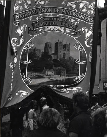 Photograph of the banner of Blackhall Lodge being carried during the Miners' Gala; the people carrying it can be seen only from the back and the banner takes up nearly all of the photograph; the banner displays a picture of Durham Cathedral, with the Fulling Mill on the River Wear in the foreground