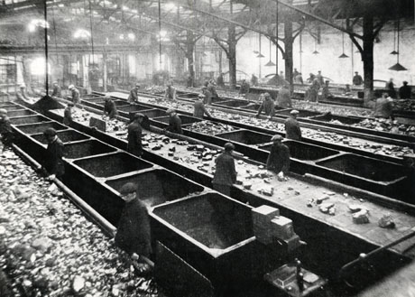 Photograph of the screens at Blackhall Colliery, run by Horden Collieries Ltd., showing the interior of a large building with a roof of cast-iron girders, from which lights are suspended; below the lights, six conveyor belts, running the length of the building, can be seen; between each conveyor belt are a line of coal trucks and at each conveyor belt are eight men inspecting the pieces of coal which cover each conveyor belt