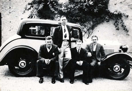 Photograph of a saloon car in two colours and with a running board, parked near a wall with a creeper growing on it; in front of the car are three men sitting on the running board and one man standing in the middle of the group and leaning against the side of the car; all four men are wearing suits but only two are wearing ties; the people have been identified as George Connell and friends, and the car as belonging to the Pieronis