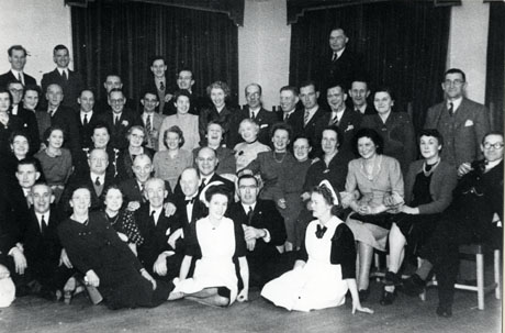 Photograph of a group of forty seven middle-aged men and women posed in front of two large curtained windows, most likely in an hotel; all of them are smartly dressed but not in evening dress; at the front of the group are two women in uniform, possibly members of staff of the hotel; at the front, is a man in a dinner jacket, who also may be a member of staff of the hotel; the group is described as Social Evening Blackhall Trades People