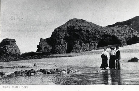 Postcard photograph entitledBlack Hall Rocks, showing the rocks jutting across the beach and , in front of them, a group of two women and a man in the dress of 1905; the dress of the figures may be seen: all three are wearing hats and all are dressed formally
