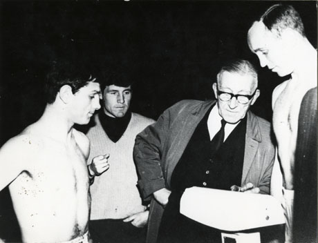 Photograph of a boxer being weighed by a middle-aged man in a raincoat and suit, while his opponent and a young man in a pullover watch.