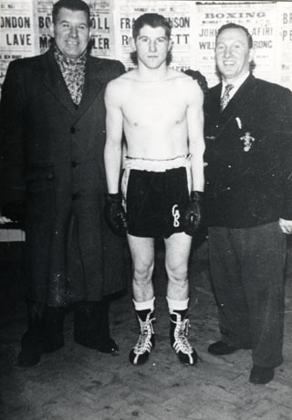 Photograph of a boxer in shorts, boxing gloves, and boots, named as George Bowes, with his father, Tom, and Terry Allen; Tom Bowes and Terry Allen are wearing an overcoat and a blazer with a nautical device on the pocket; all three are photographed against a background of boxing posters