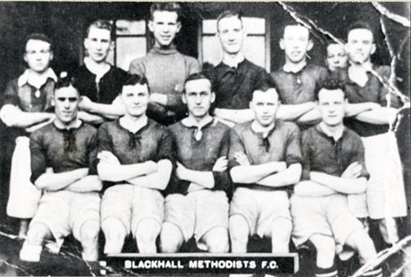 Photograph showing eleven men in football strip with the face of a twelfth man peering over the shoulders of the two men on the right of the back row; they have been identified as the members of Blackhall Methodists Football Club with their coach