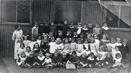 Photograph of a group of fifty four small children, aged between approximately five years and seven years, taken outside a building of corrugated iron, accompanied by a woman, presumably a female teacher; in front of the children is a board, possibly bearing the words Blackhall Colliery School; the photograph is described as 1st School in Blackhall, 1917