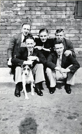 Photograph of a group of five men and a dog taken against a brick wall , described as Group of Friends; the men are all wearing suits and ties and squatting down