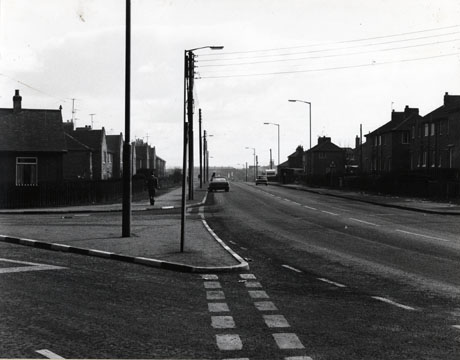 Photograph described as Road to Blackhall showing a road lined with housing of the 1950s and the junction of a side road with it ; the road is clear, apart from a car parked on the left-hand-side of the road and a car driving down the right-hand-side of the road; one man can be seen in the middle distance walking away from the camera, and a figure can be seen behind the parked car.