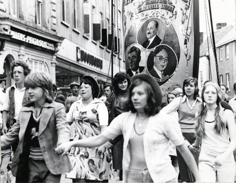 Photograph showing the Blackhall Lodge banner being carried past Woolworth's in The Market Place, Durham City; the people carrying the banner cannot be seen as they are obscured by a group of young people in front of the banner; the banner can be seen and shows the portraits of Earl Attlee, Aneurin Bevan, and Arthur Horner.