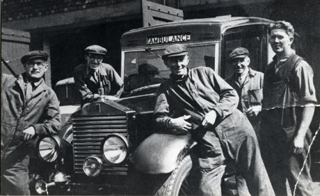 Photograph showing what have been identified as the garages at Blackhall Colliery; in the background the open doors of the garages may be seen and the indistinct image of a vehicle; the majority of the photograph shows five men in overalls standing next to, and leaning on, a, possibly, Rolls-Royce vehicle with the word Ambulance across its top.