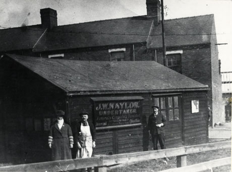 Photograph of the exterior of a one-storeyed wooden building with, behind it, the upper windows and roofs of three terraced houses; on the side of the wooden building is a sign reading, as follows: J. W. Naylor, Undertaker. Funerals Completely Furnished. Distance No Object. Attendance Day and Night Joiner and Cabinet Maker; in front of the building is woman in a coat, hat and scarf, a man wearing a workman's apron over a suit, and a second man wearing a working suit; they have been identifed as being in Blackhall
