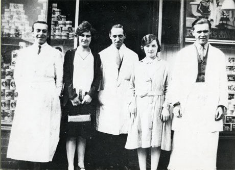 Photograph of the staff of Thompson's Red Stamp Store, Blackhall, photographed outside the shop; the photograph shows two men wearing white coats and aprons, one man wearing a white overall, one woman wearing an overall, and one woman dressed in normal indoor clothes; behind the group, the windows of the shop may be seen containing pyramids of tins and an indistinct advertisement for bath cubes.