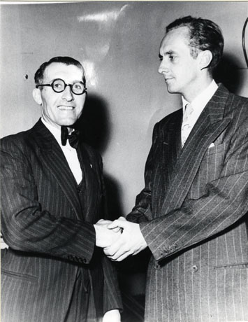 Photograph of Tom Hughes on the left, shaking hands with Hughie Green, on the right; both men are wearing pin-striped suits, but the occasion is unidentified and only the men may be seen.