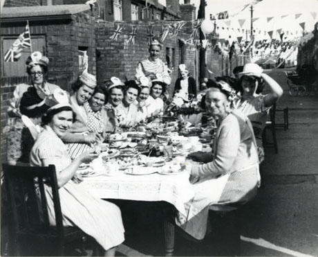 Photograph of approximately fourteen women and one man seated round a table, identified as being in the back lane at Meadow Avenue, Blackhall, to celebrate the Coronation of Queen Elizabeth II on 6 June 1953; the people are sitting round a table covered with a cloth on which plates of food are standing; all the people are wearing fancy hats and Union Jacks are strung across the road.