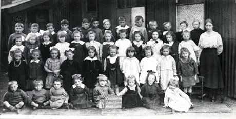 Photograph of a group of forty three children, aged approximately six years, and a woman, presumably their female teacher, outside the Tin School in Blackhall; at the front of the group is a board bearing the words Blackhall Coly. Infant School Class 2 1919