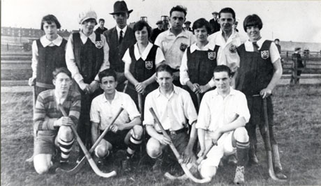 Photograph of six male and five female hockey players posed with a middle-aged man in a field with, in the far distance, rows of houses and the colliery wheels; closer behind the group, the heads of seven boys wearing flat caps may be seen observing the group; the members of the group are wearing a badge bearing crossed hockey sticks and, possibly, the letters B H C (possibly Blackhall Hockey Club); the women are wearing gym slips and the men shirts, shorts and long socks.