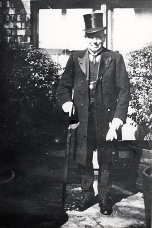 Photograph, described as being of Mr. Naylor, First undertaker in Blackhall, showing him dressed in a frock coat, wearing a top hat, and carrying a rolled umbrella; he is photographed outside what appears to be a house.