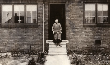 Photograph of the exterior of a brick bungalow with steps leading to the doorway and windows of three casements either side of the door; a woman, wearing an apron and a dark skirt, is standing on the steps; the photograph is taken fairly close-up and details of her clothing may be seen; the scene has been identified as being in Blackhall