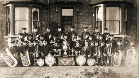 Photograph of the twenty seven members of Blackhall Colliery Band posed outside a house with two bay windows; all but one of the men of the band are wearing the uniform of the band; all are holding their instruments and the trophies won by the band are displayed in front of them