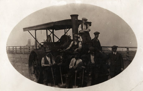 Photograph, entitled Building the Coast Road, showing nine work men standing in front of, and sitting and standing on, a steam roller; in the distance the pit wheels of, possibly, Blackhall Colliery may be seen; the men and the roller may be seen in the foreground