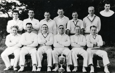 Photograph of the members of Blackhall Welfare Cricket Club Team, showing twelve men in cricketing whites and a trophy cup on the ground in front of them; the head and shoulders of a thirteenth man may be seen in the top right-hand corner of the photograph