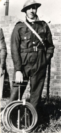 Photograph of R. Turnbull, Air Raid Warden at Blackhall Colliery during the Second World War, showing Mr. Turnbull full-length, close-up, wearing A.R.P. overalls, a tin hat, and holding a stirrup pump