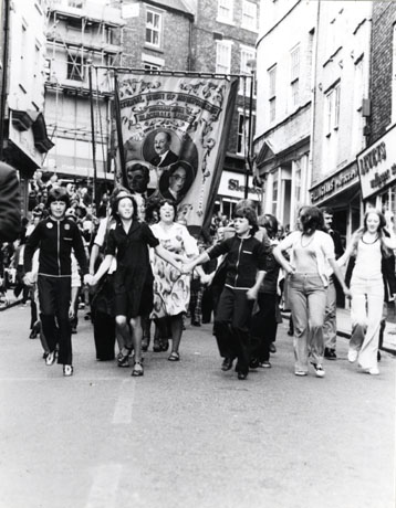 Photograph of the Blackhall Lodge banner being carried down Elvet Bridge in Durham City during the Miners' Gala; in front of the banner are a number of young people holding hands; the banner shows Earl Attlee, Aneurin Bevan and Arthur Horner