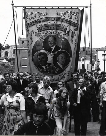 Photograph of the Blackhall Lodge banner being carried along Framwellgate Bridge in Durham City, during the Miners' Gala; the lodge officials may be seen, with a number of young people; the banner shows portraits of Earl Attlee, Aneurin Bevan and Arthur Horner