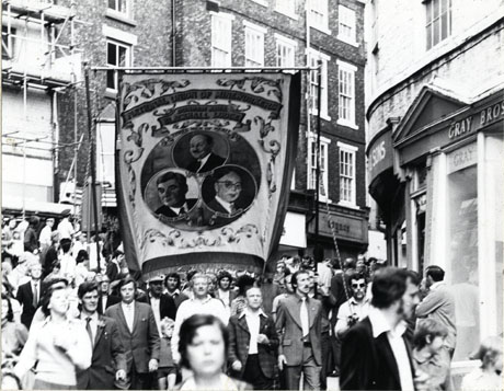Photograph of the Blackhall Lodge banner being carried along the top of Elvet Bridge in Durham City, near Saddler Street, with the lodge officials and crowds surrounding them; the banner shows portraits of Earl Attlee, Aneurin Bevan and Arthur Horner
