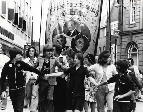 Photograph of the Blackhall Lodge banner being carried through Durham Market Place at the top of Silver Street, during the Miners' Gala; in front of the banner are a number of young people holding hands ; the banner shows portraits of Earl Attlee, Aneurin Bevan and Arthur Horner