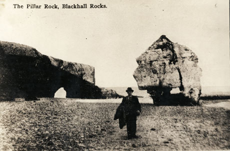 Postcard photograph entitled The Pillar Rock, Blackhall Rocks, showing the pillar rock and another rock jutting into the sea with a cave in it; in the foreground is the indistinct figure of a man wearing a suit, Trilby hat and carrying a coat over his arm; the photograph is described as Much-Upon-Little
