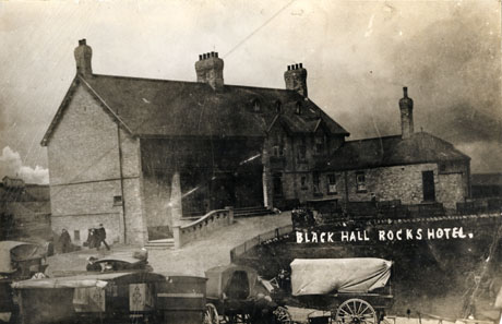 Photograph of the exterior of Blackhall Rocks Hotel showing the facade of the hotel, six horse-drawn vehicles in the foreground of the picture, and three indistinct male figures walking down steps at the side of the hotel away from the camera and carrying cases; a number of very indistinct figures may be seen near some seats at the front of the hotel