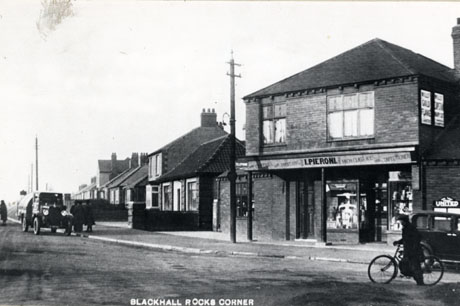 Postcard photograph entitled Blackhall Rocks Corner, showing a street with houses receding into the distance and, in the foreground, the facade of a shop, above which is written: Groceries and Provisions I. Pieroni High Class Ices and Confectionery; a vehicle, possibly a dust cart, is parked at the side of the road and, standing by it, are three indistinct men; on the right of the photograph a man on a bicycle and the back of a car may be seen: advertisements for Gold Flake cigarettes and for the United Bus Company can be seen indistinctly on the side of the shop