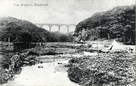 Postcard photograph entitled The Viaduct, Blackhall, showing, in the distance, the viaduct; in the foreground a stream, trees, and grass may be seen; to the right of the picture, in the middle ground, a small pointed tent may be seen, with an indistinct figure beside it; the photograph is also described as Boy Scouts Camping at Deneholme