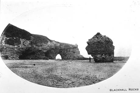 Postcard photograph, entitled Blackhall Rocks, showing the cliff rock with a hole in it and the detached rock at Blackhall; a small indistinct figure can be seen near the detached rock; the foreground of the photograph shows the beach