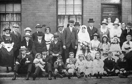Photograph showing twelve men, fifteen women, and nine children standing in three rows against the wall of a house; the children, and two men, are sitting on the edge of the pavement; the bride in her wedding dress and the groom, are standing surrounded by the other people, all of whom are dressed formally in suits and dresses and hats; they have been identified as Joe Rodger's Wedding, Blackhall