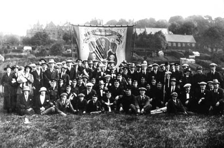 Photograph showing a group of approximately eighty people, and bandsmen in uniform, grouped in front of a banner of the Durham Miners' Association, showing the portraits of three men; behind the group, are allotments on the right of the picture and, on the left, a large elaborate building; the photograph has been identified as being at Blackhall